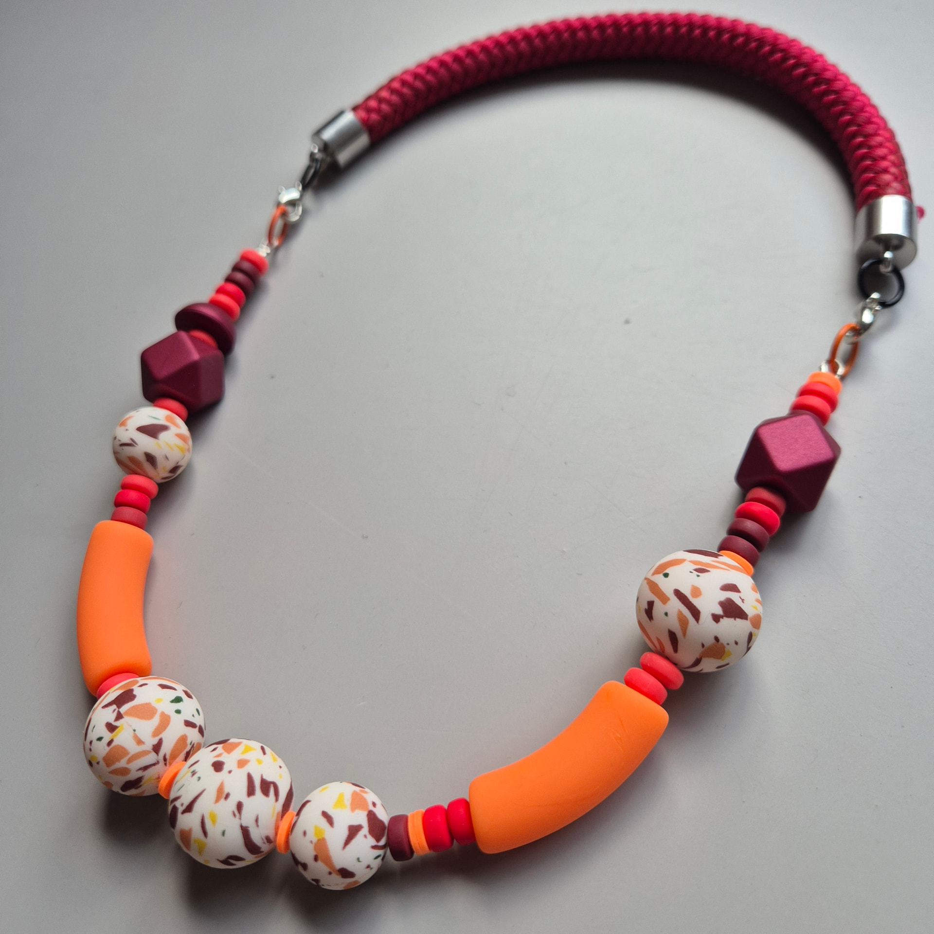Necklace of white terrazzo spheres, orange curved tubes and burgundy geometric beads on a burgundy braided rope