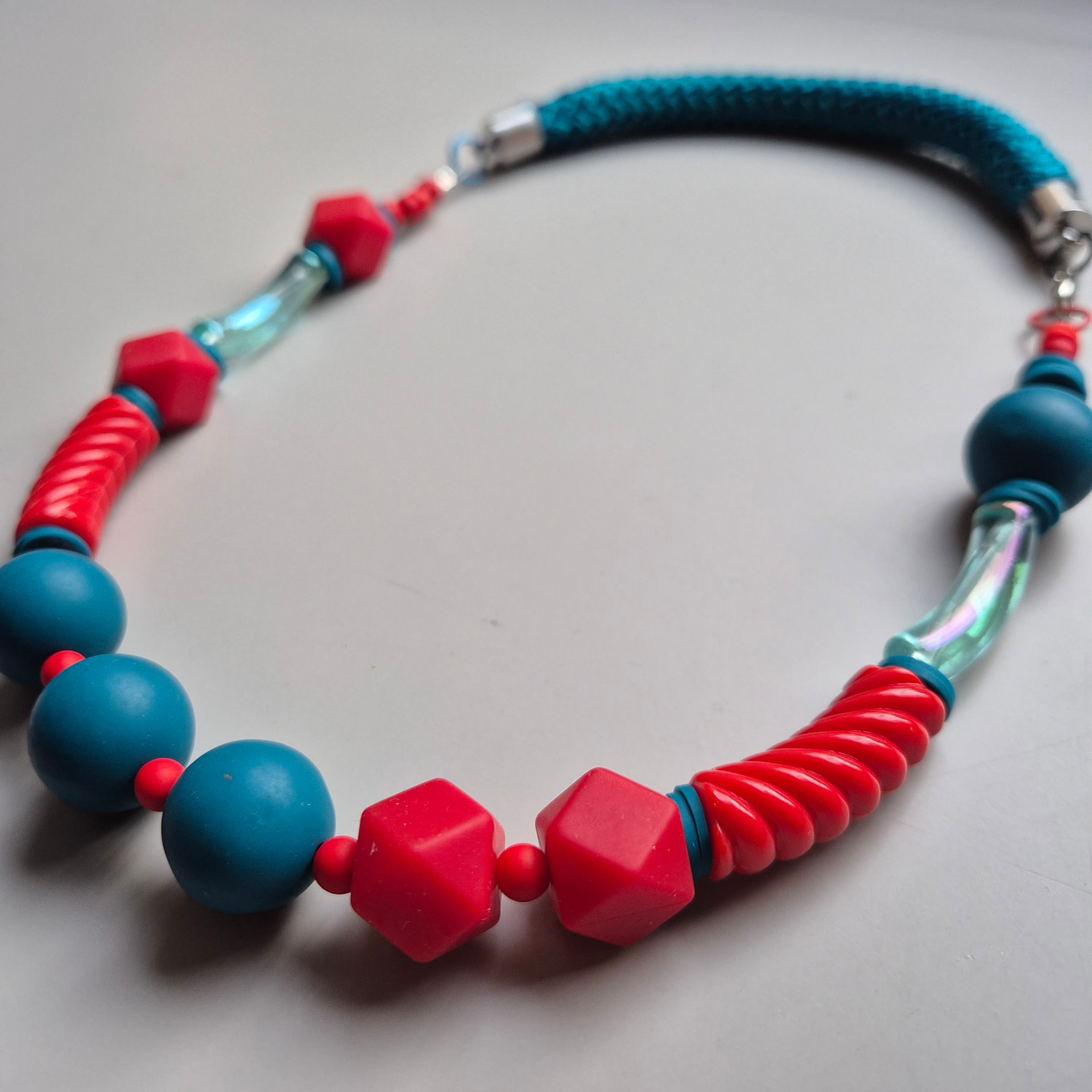 Necklace of red faceted beads, ribbed red tubes, teal spheres and iridescent glass on a teal braided rope