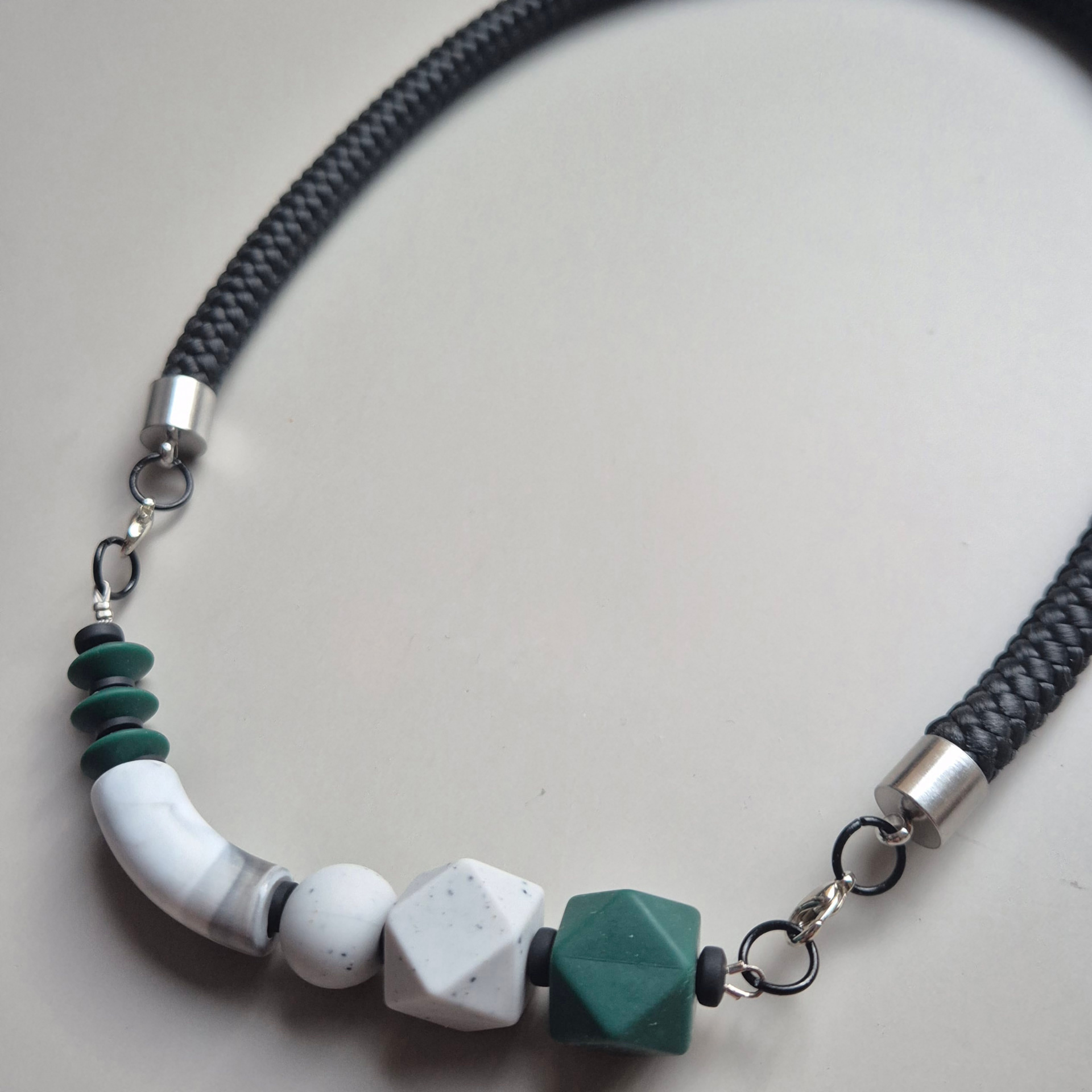 Close detail of the necklace with green discs, a white marbled curved tube, speckled grey sphere and facet, green geometric bead and silver clasp on a black rope