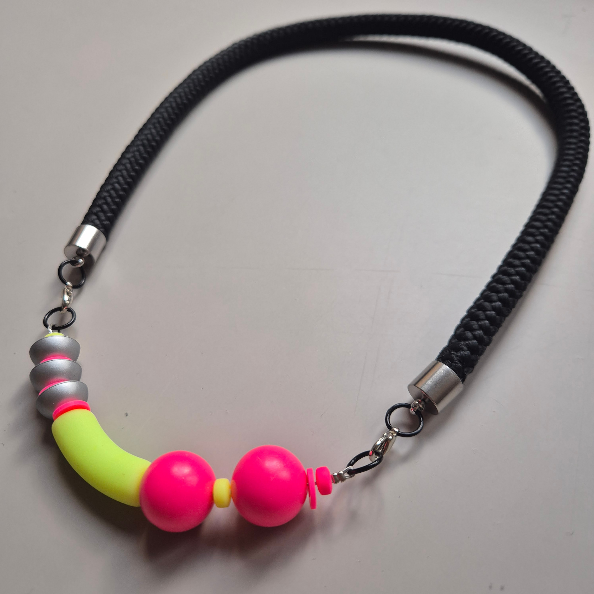 Necklace with neon pink spheres, a neon yellow curved tube and silver ribbed discs on a black rope
