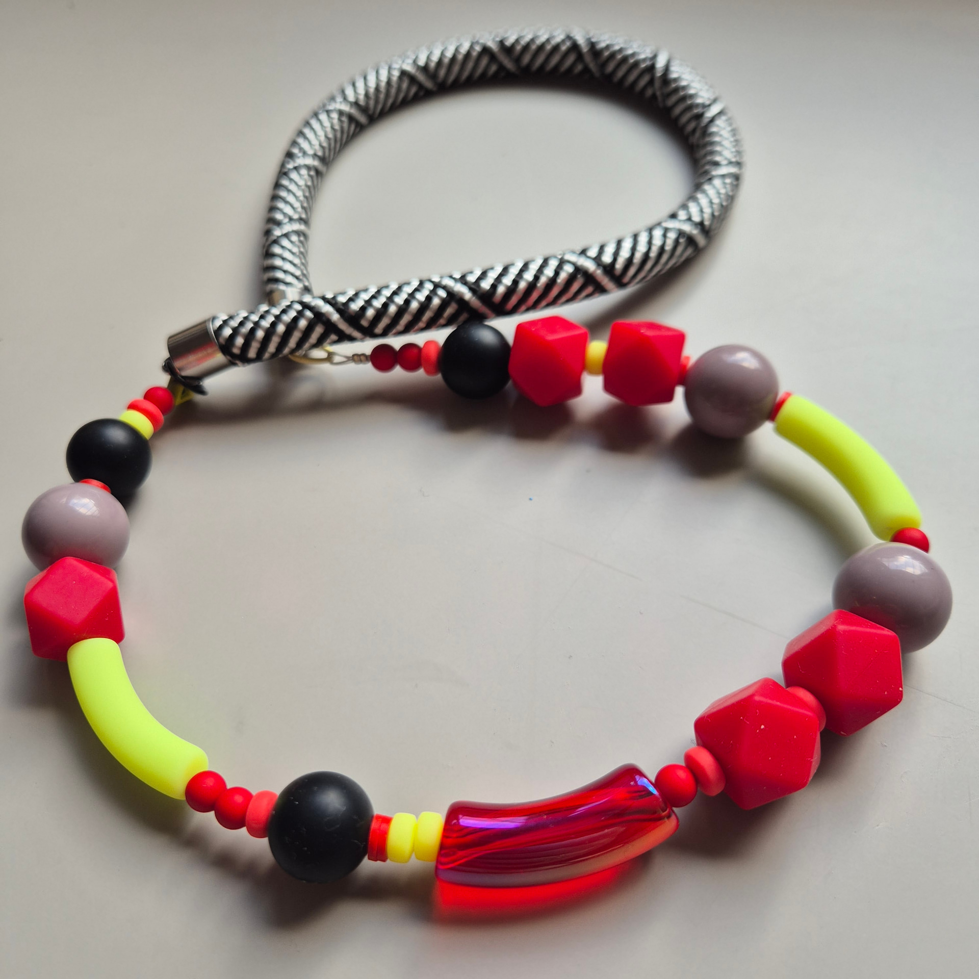Necklace with red geometric facets, neon yellow tubes, black spheres and an iridescent red curved tube on a black-and-white rope