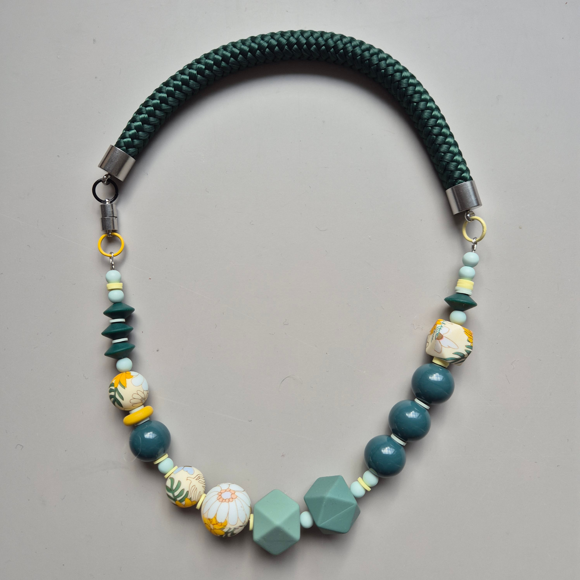 Full view of the Jardin 2.0 necklace with floral beads, sage hexagons and deep teal spheres on a dark green braided rope