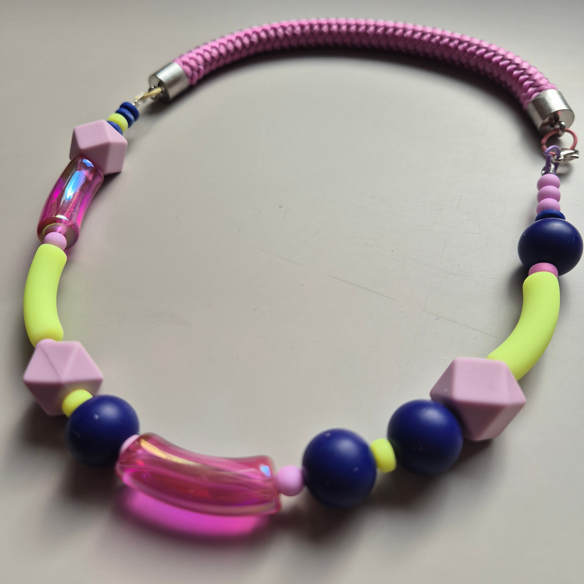 Necklace with navy blue spheres, neon yellow tubes, iridescent pink curved tubes and pink faceted beads on a pink braided rope